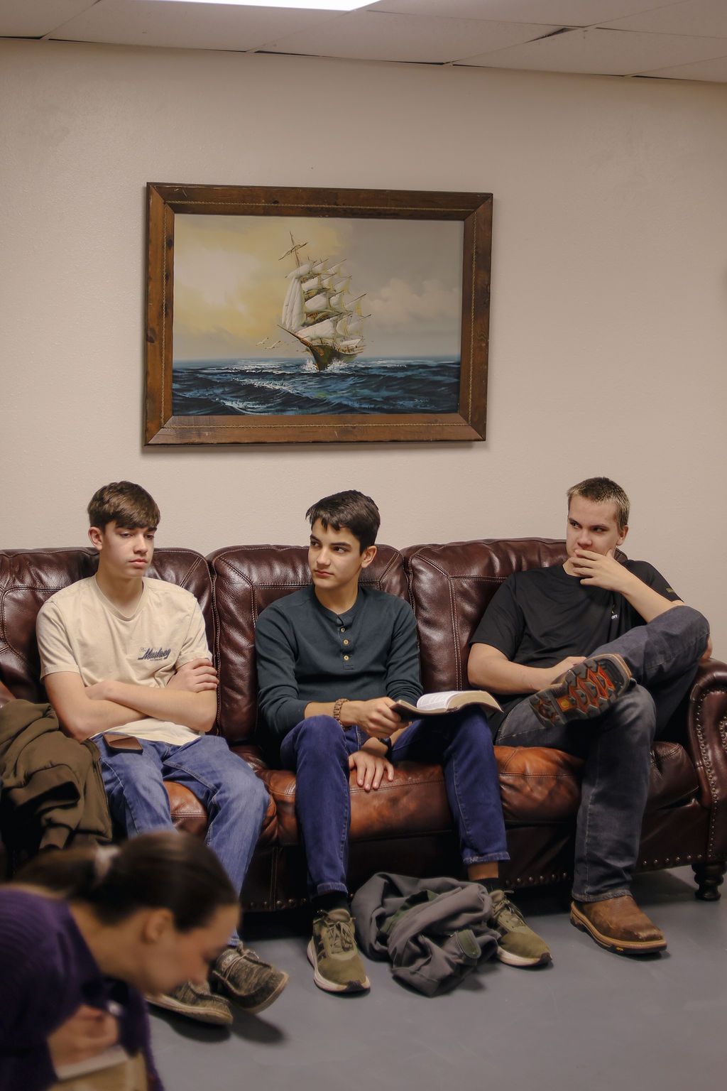 Three people sit on a brown leather couch in a room with a framed ship painting on the wall.