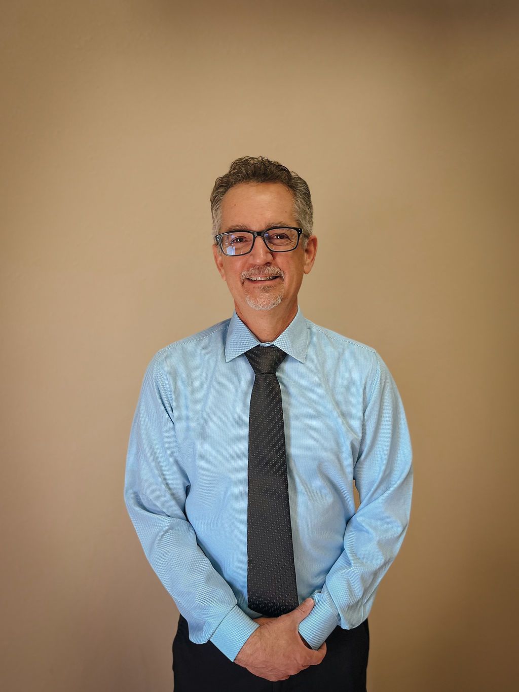 A person wearing glasses, a light blue long-sleeved shirt, and a dark patterned tie stands against a plain tan wall.