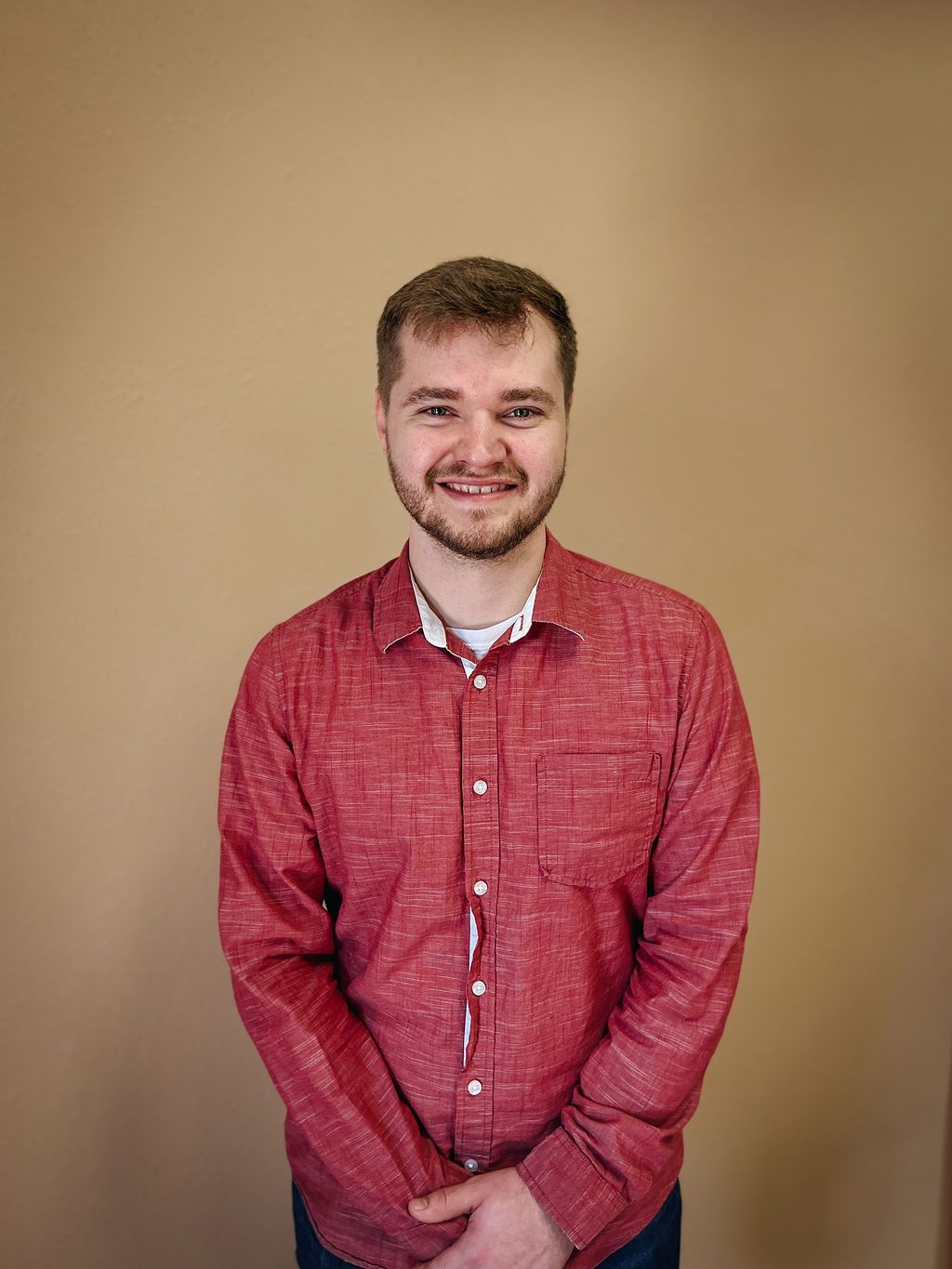 A smiling person with a beard stands against a plain beige wall, wearing a textured red button-down shirt.