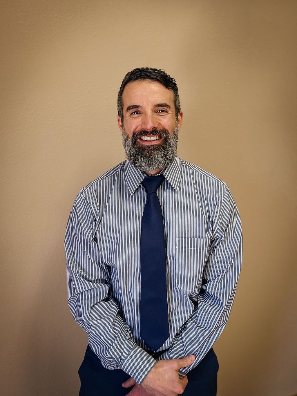 A smiling person with a beard wearing a blue and white striped shirt and a solid dark blue tie against a tan background.