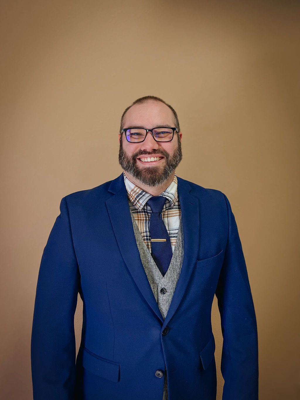 A smiling person with a beard and glasses, wearing a blue blazer, patterned shirt, grey vest, and blue tie against a wall.