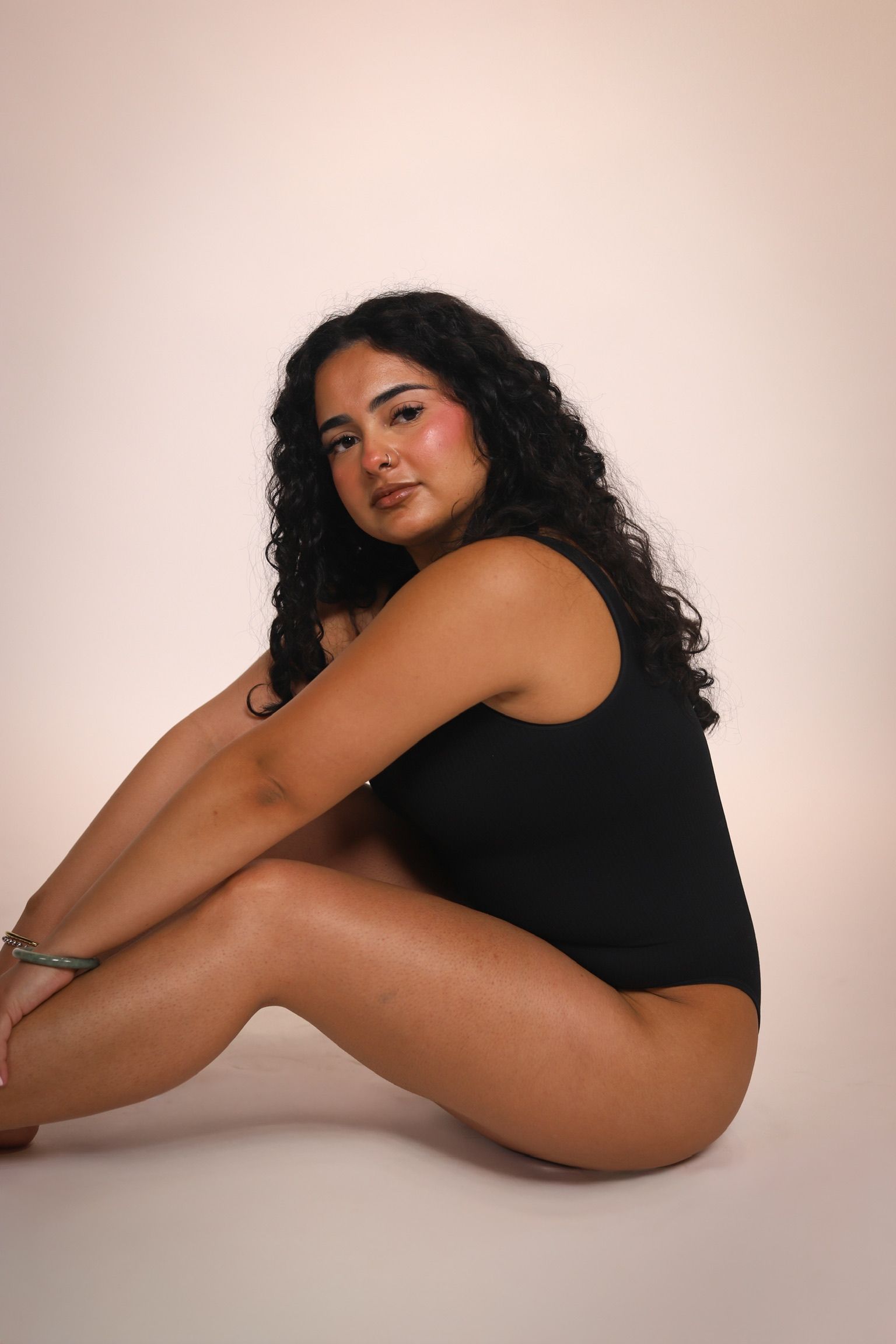 Seated woman in a black outfit on a light studio backdrop, posed with knees bent and a calm expression