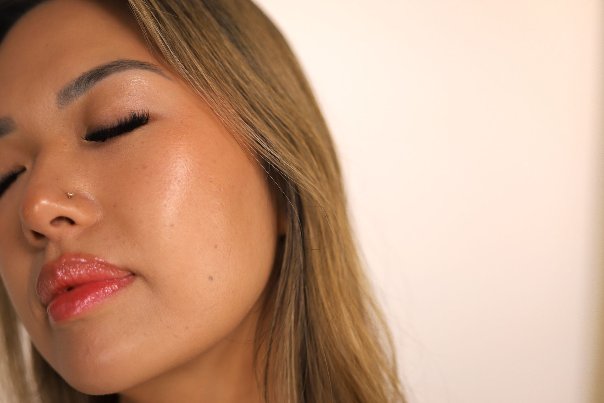 Close-up of a woman with eyes closed, wearing glossy pink lipstick against a soft beige background