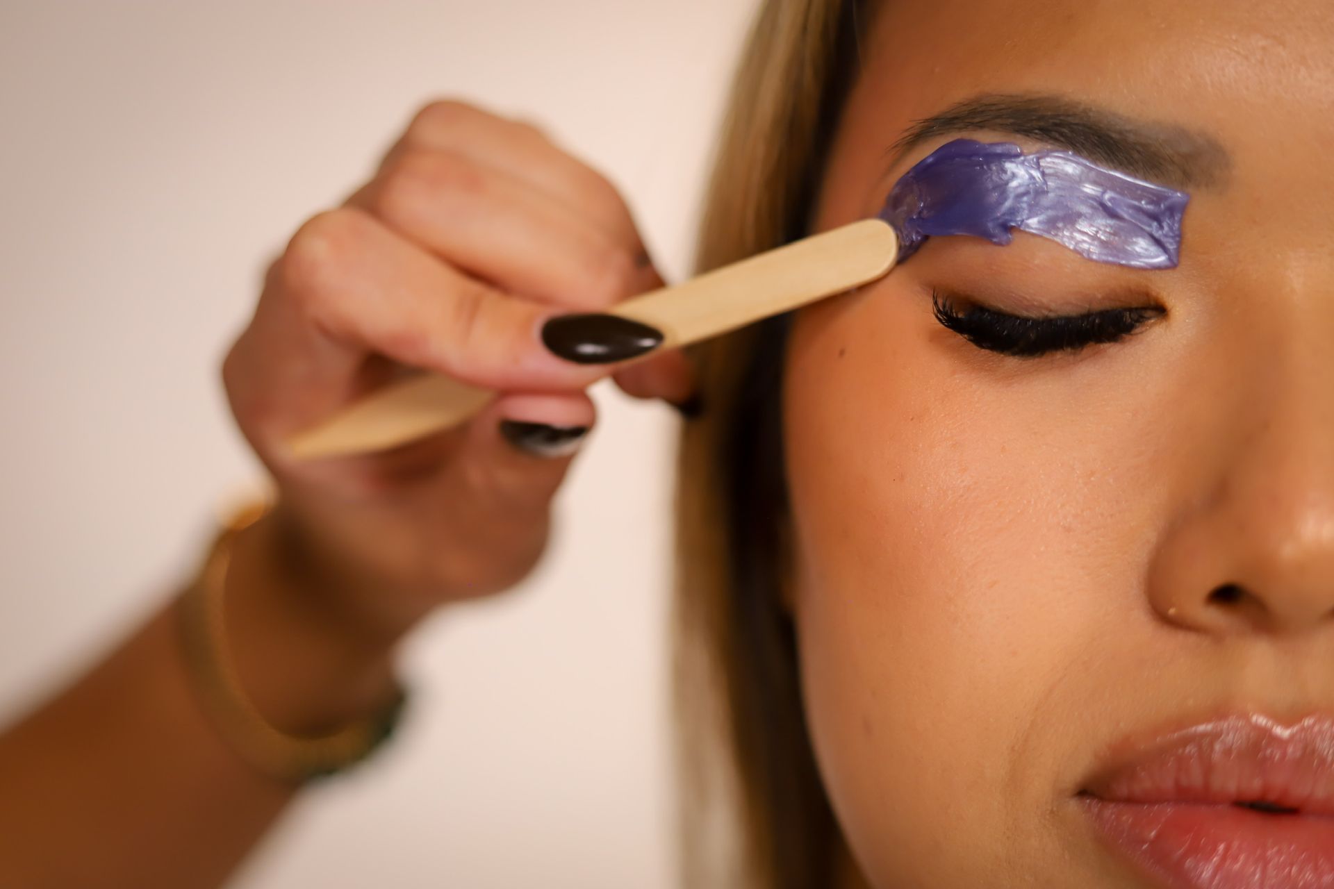 Woman's eyebrow being waxed; pink wax applied by a gloved hand with a wooden applicator.