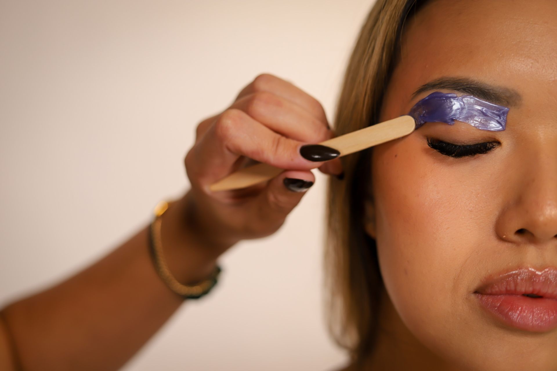 Woman's eyebrow being waxed; pink wax applied by a gloved hand with a wooden applicator.