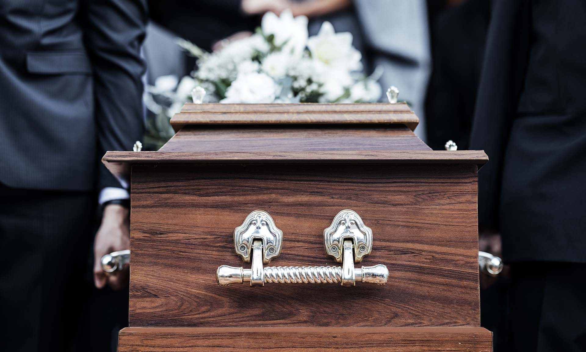 People carrying a wooden casket decorated with white flowers, likely at a funeral.