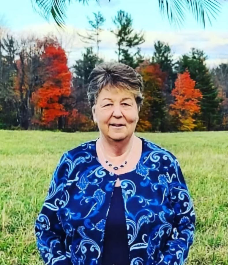 A woman in a blue jacket is standing in a field with trees in the background.