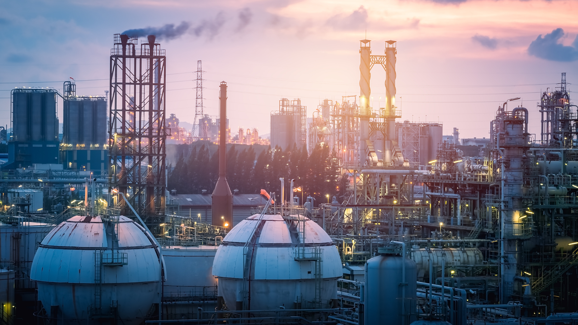 Industrial plant at sunset, with tanks, smokestacks, and pipes against a colorful sky.