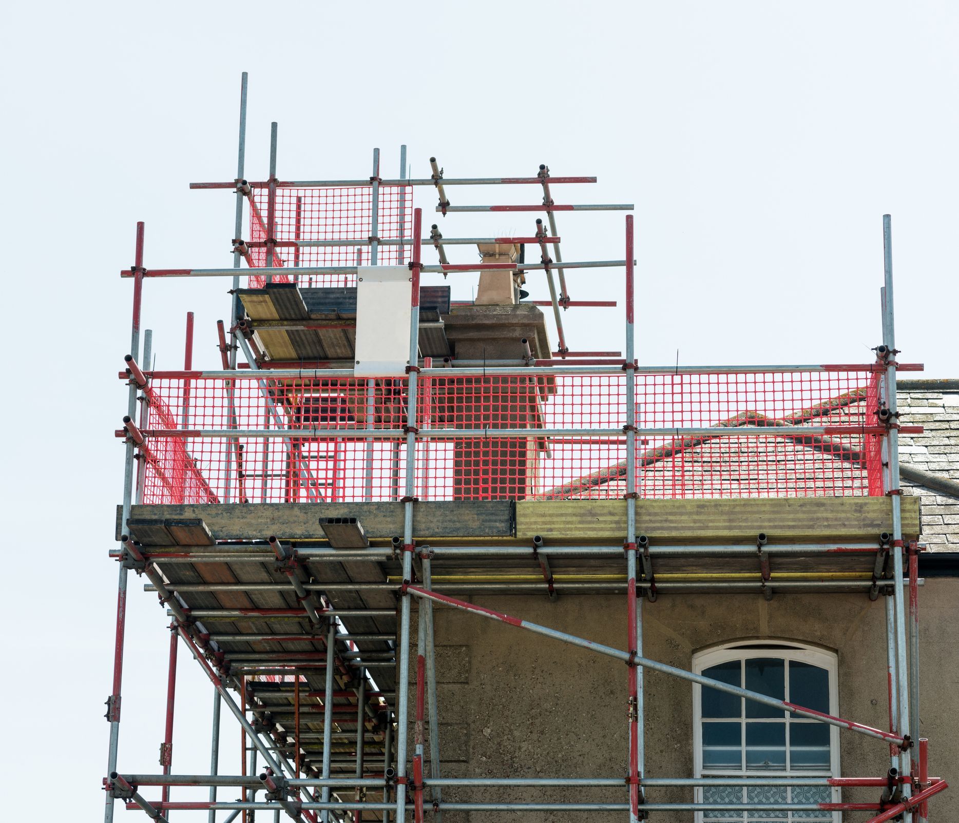Scaffolding around a building's roof for construction work. Red safety netting and grey building.