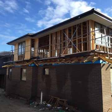 Construction site: partially built two-story house with exposed wooden framing and brick exterior under a blue sky.
