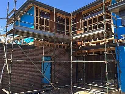 Scaffolding surrounds a two-story brick house under construction; blue tarp covers some walls.