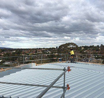 Person working on a corrugated metal roof, wearing a hard hat. Safety railings in foreground, houses and sky in the background.