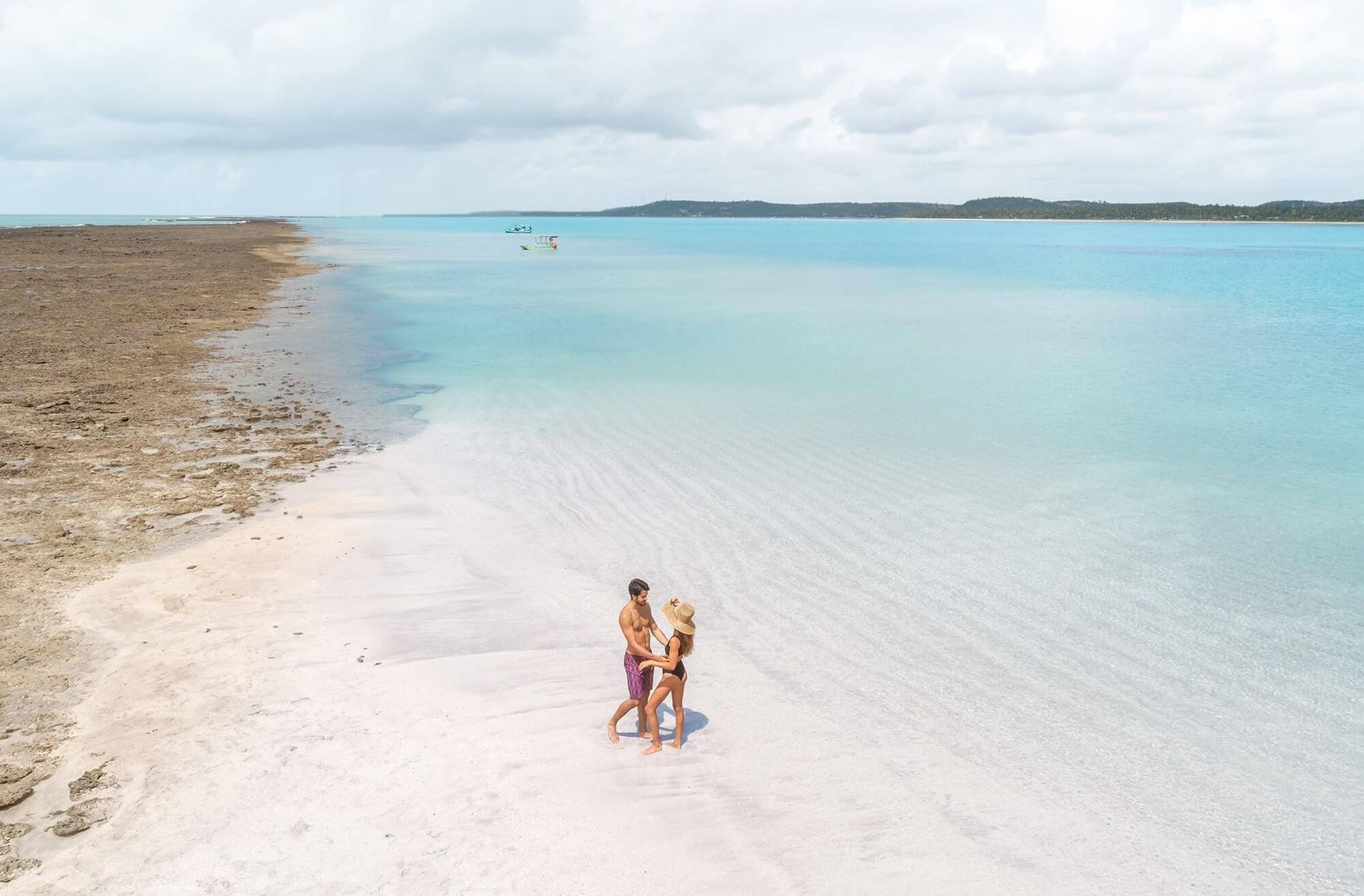 Duas pessoas em pé numa praia de areia branca junto à água cristalina turquesa.