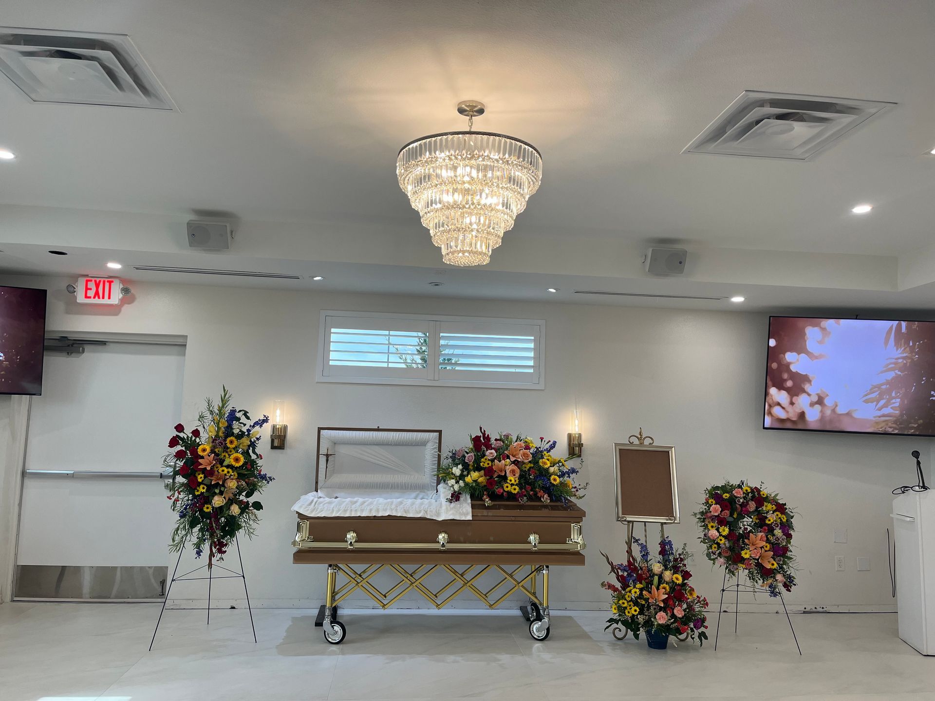 Funeral chapel with a casket centered beneath a chandelier, surrounded by floral wreaths and arrangements.