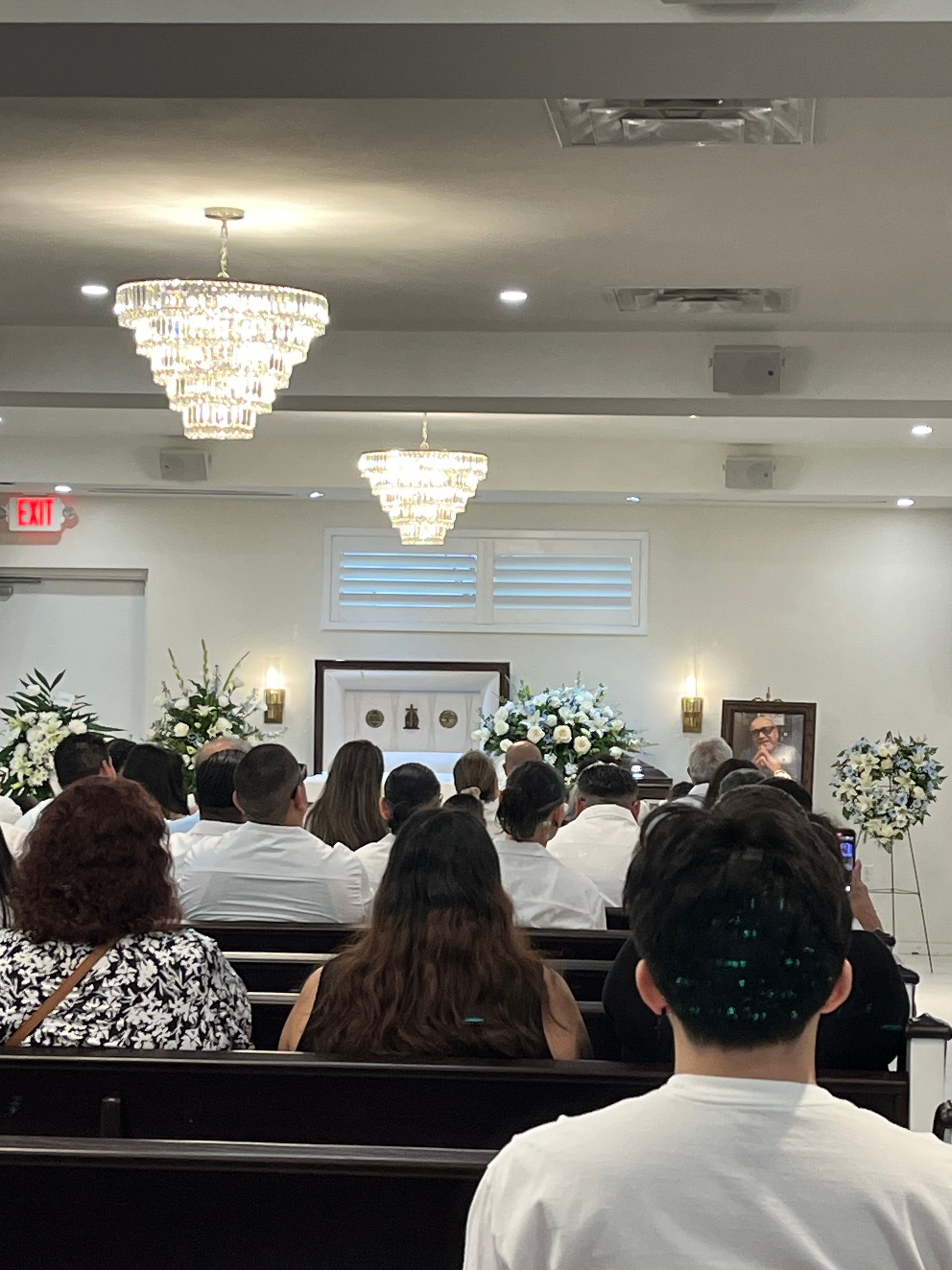 Mourners seated in a funeral home chapel facing a casket and floral arrangements during a memorial service
