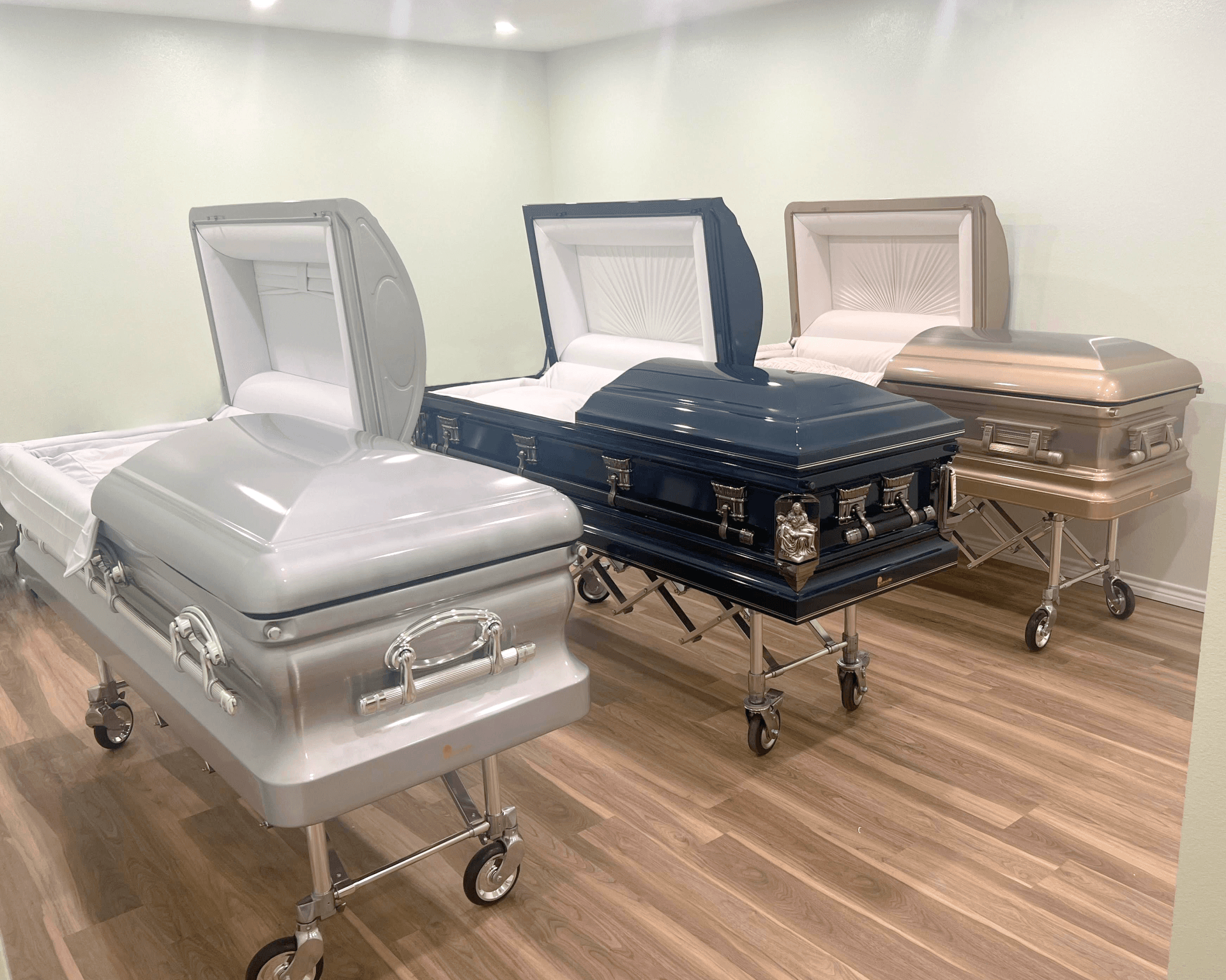Three open caskets in a funeral home showroom, in silver, black, and beige finishes.