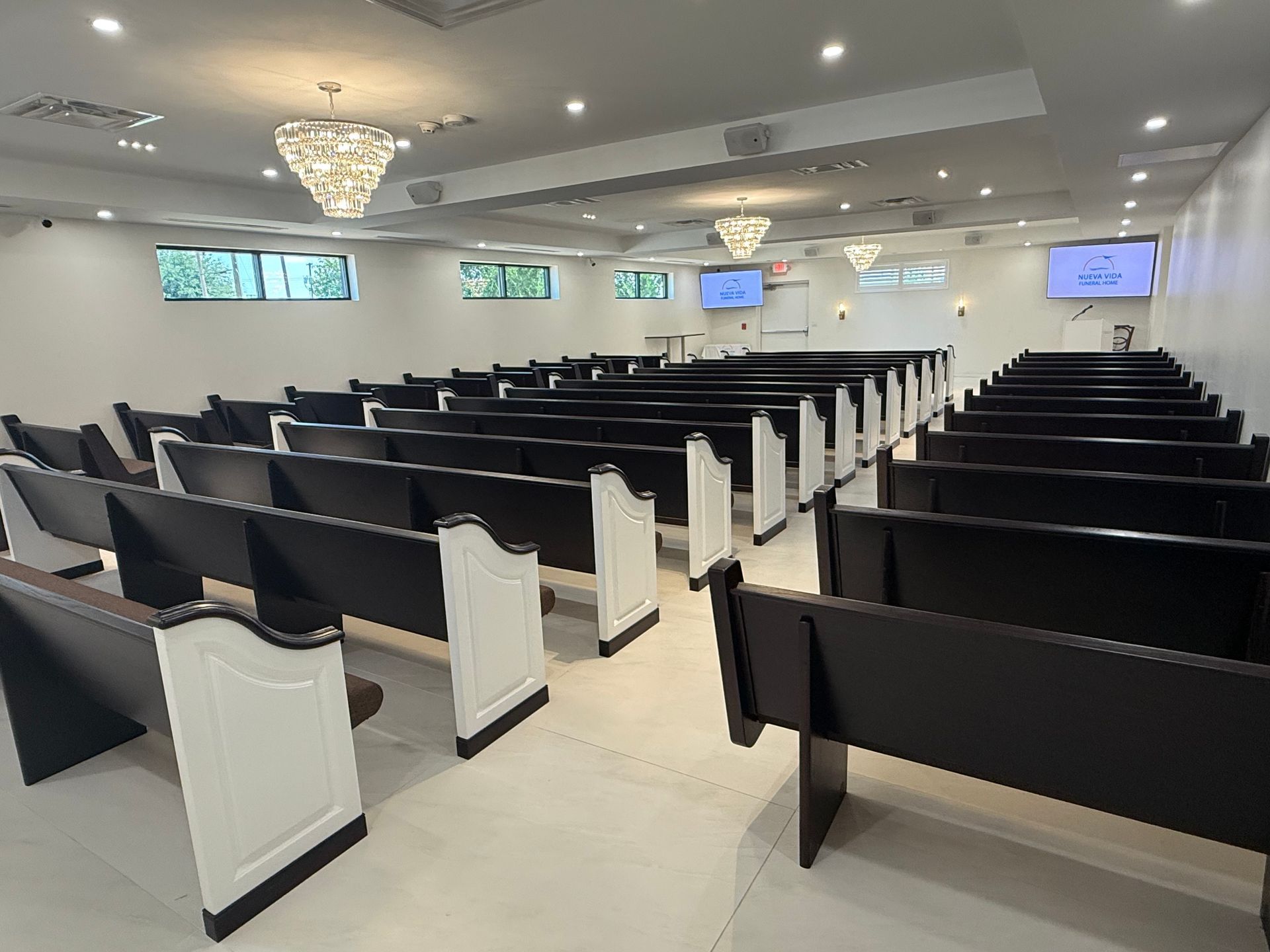 Empty modern auditorium with rows of black and white benches, chandeliers, and wall-mounted screens.