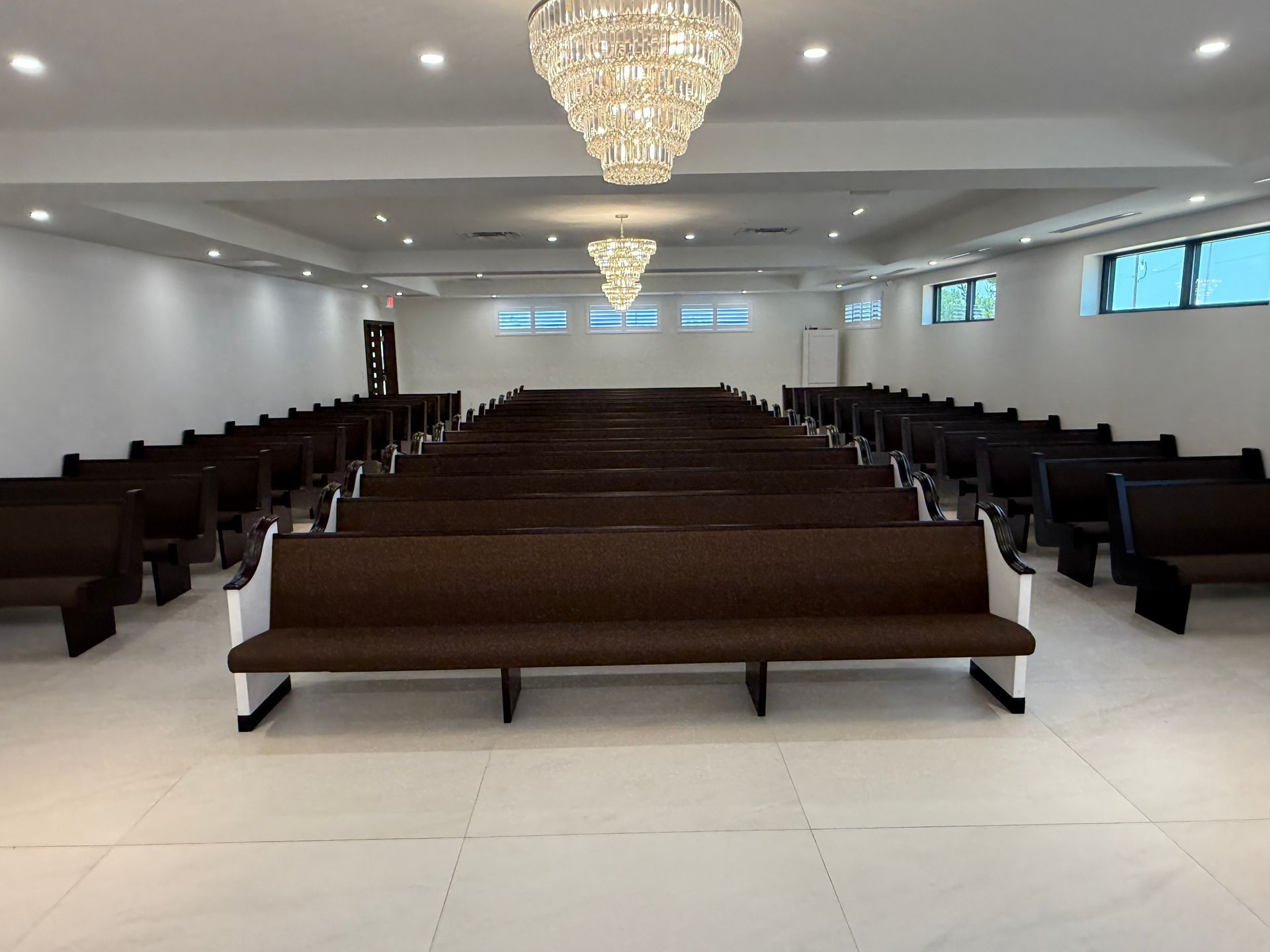 Empty modern chapel with rows of dark pews, white floor, and crystal chandeliers under a high ceiling