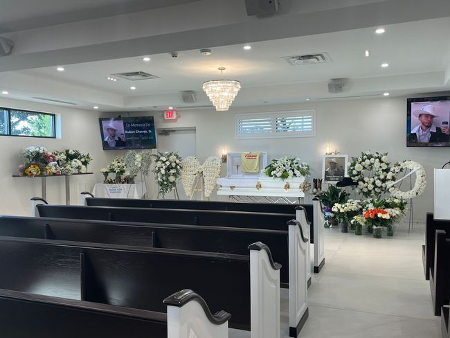 Funeral chapel with white flower arrangements, rows of black chairs, and casket at the front.