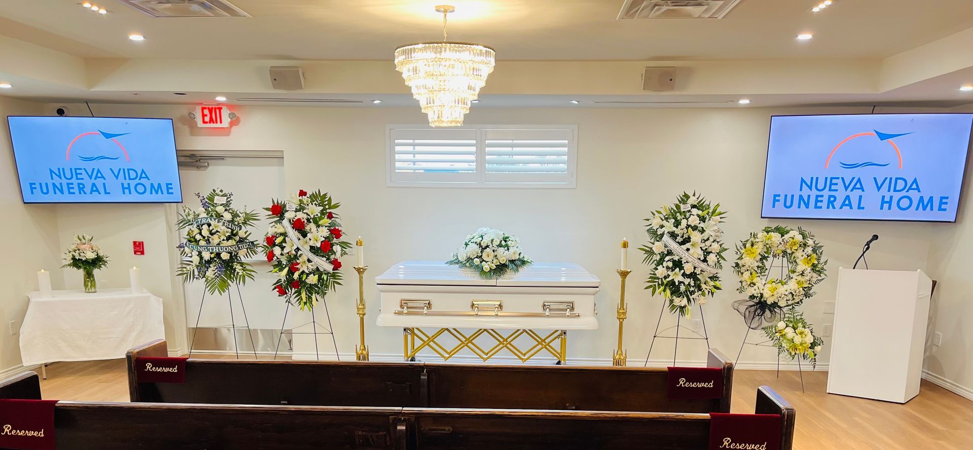 White funeral home chapel with casket, flowers, chandeliers, and two blue screens at the front.