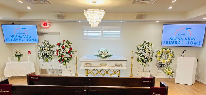 White funeral home chapel with casket, flowers, chandeliers, and two blue screens at the front.