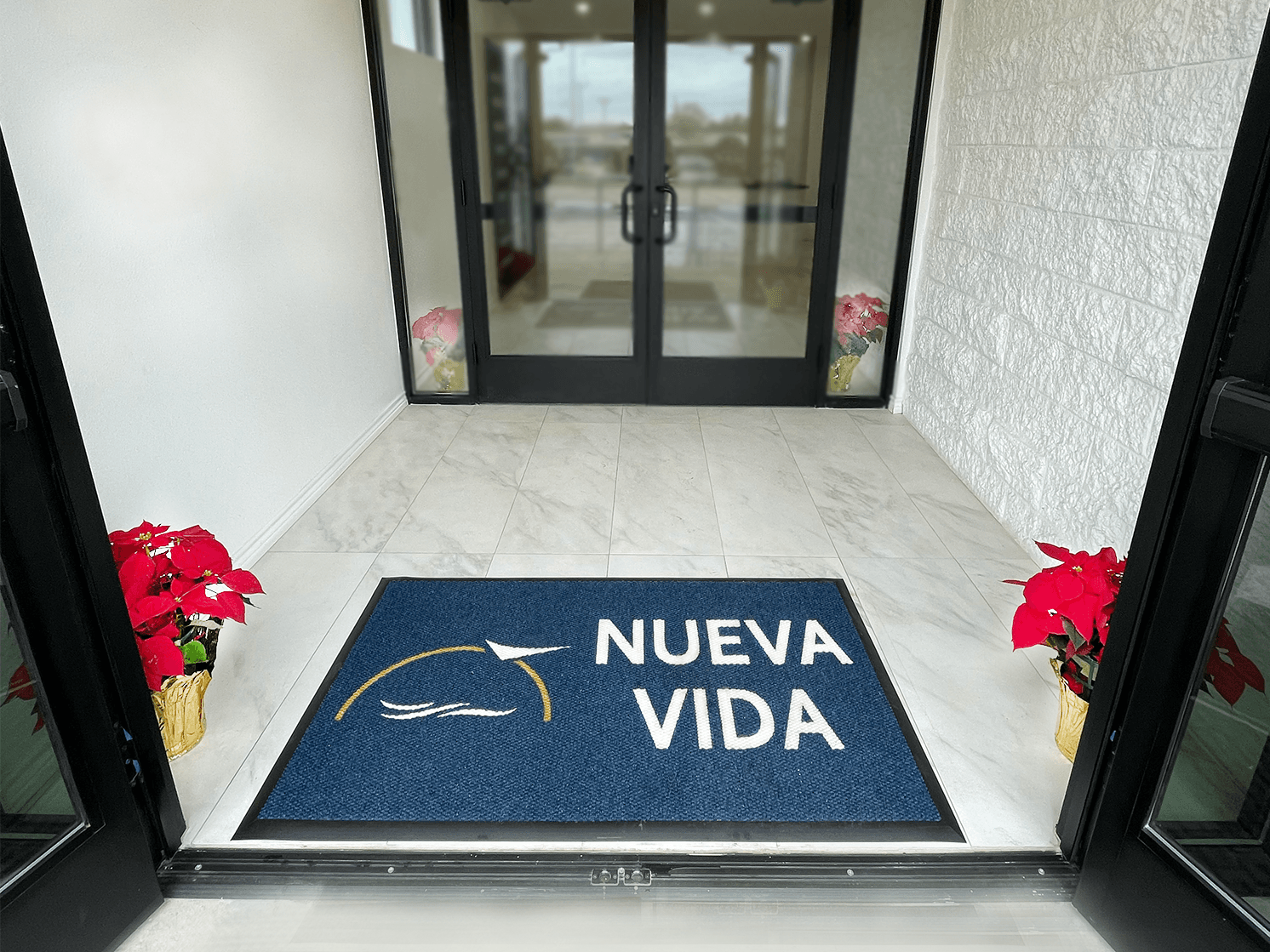 Entrance with blue “NUEVA VIDA” doormat, glass double doors, and red potted flowers on either side.