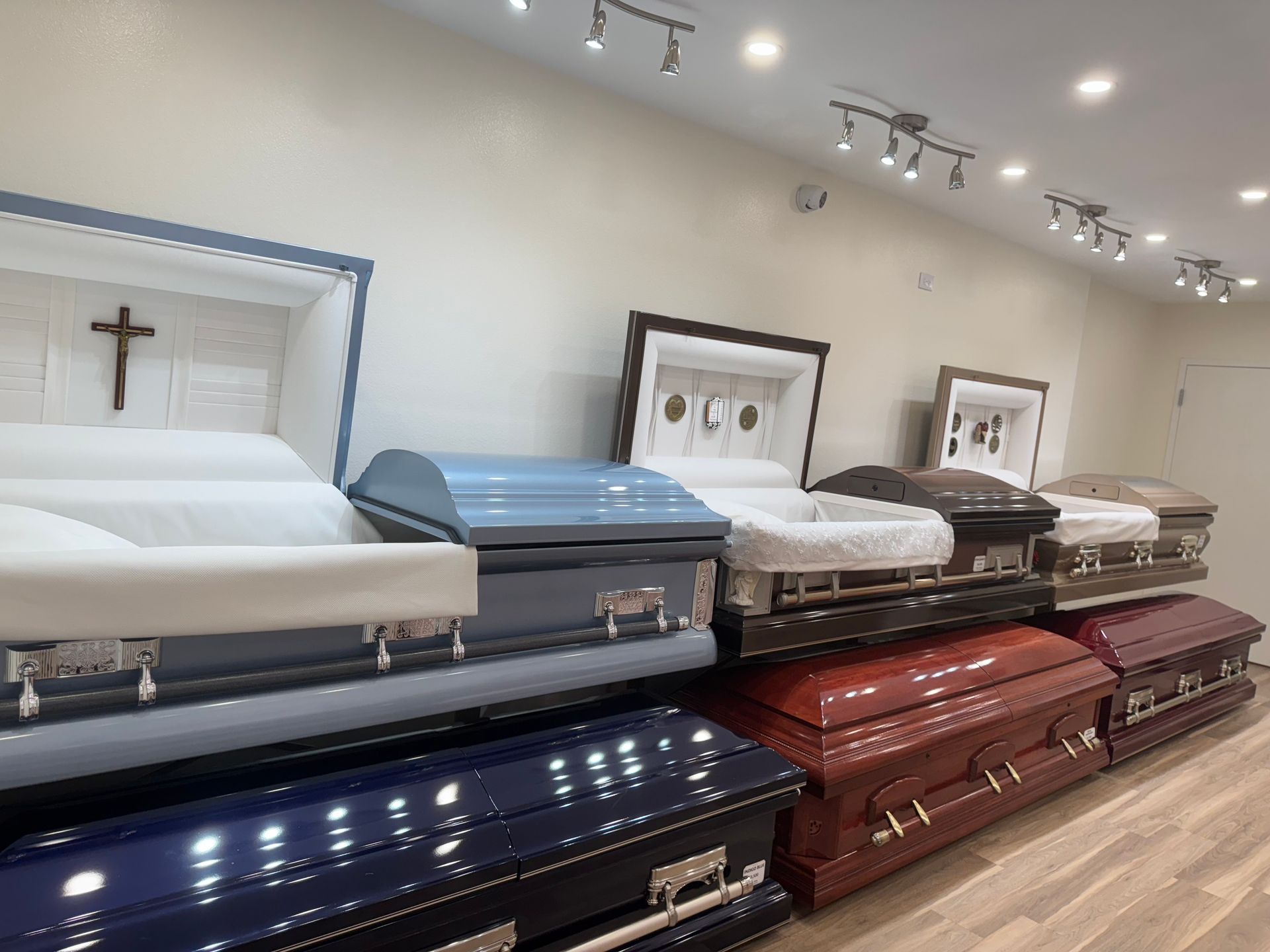 Funeral home showroom with several open caskets in white, blue, brown, and burgundy under track lighting