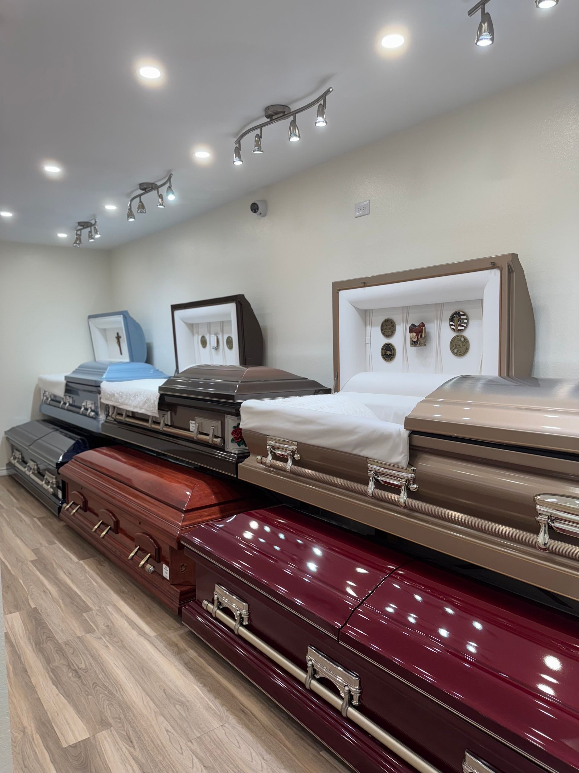 Rows of coffins in a funeral showroom with white walls, track lights, and wood flooring