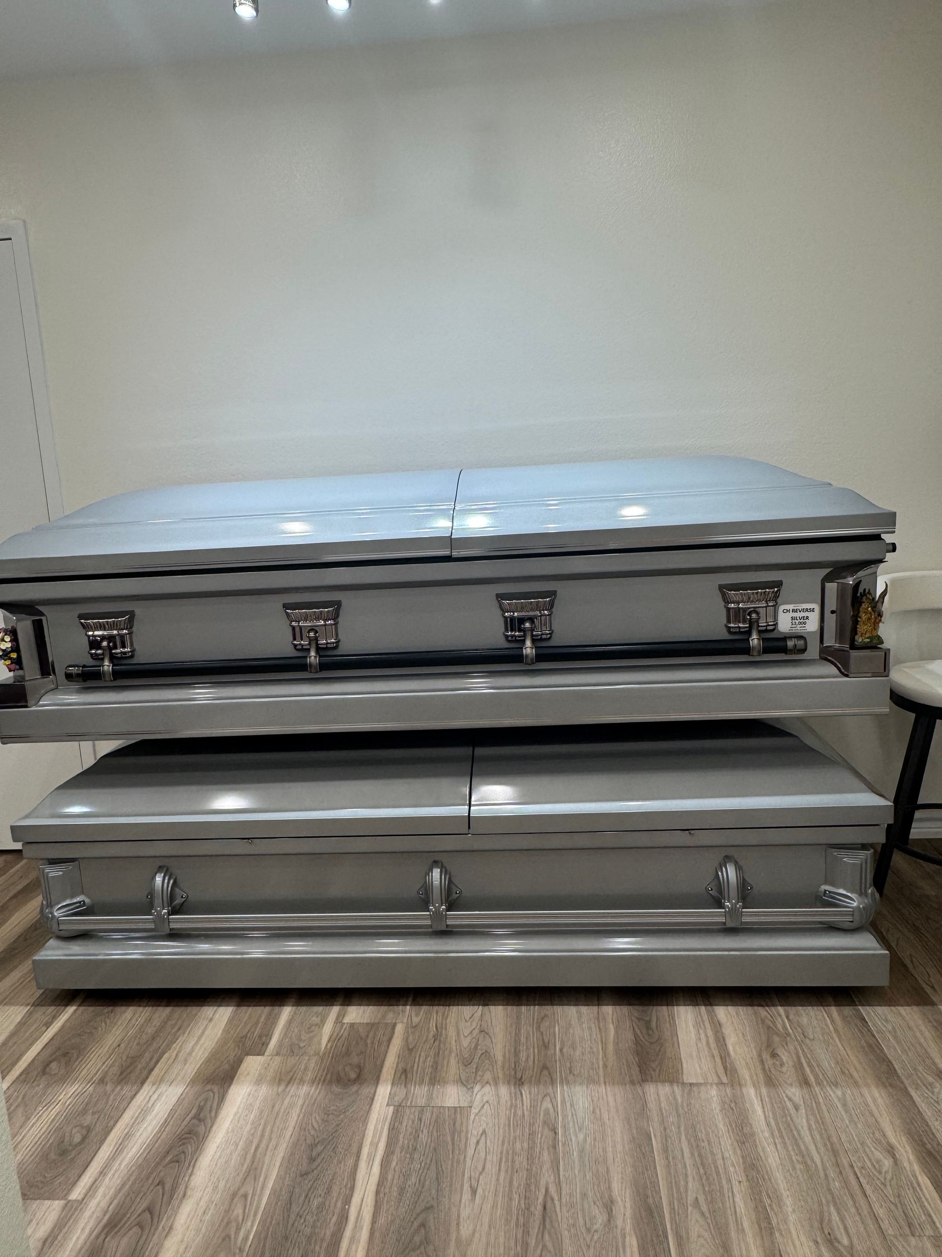 Two silver caskets stacked in a room with beige walls and wood-patterned flooring