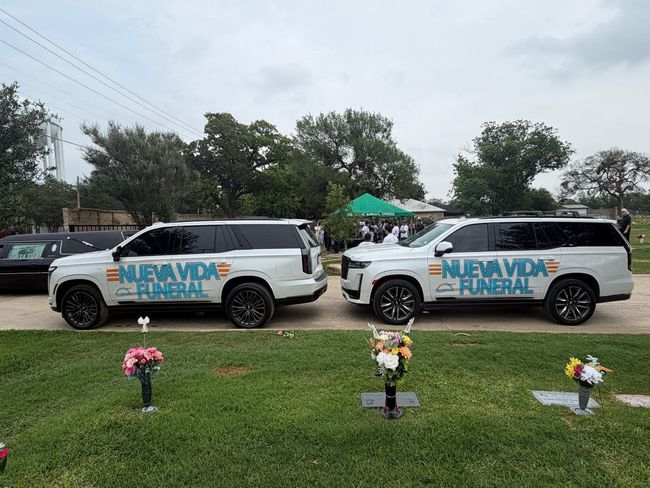 Two white Nueva Vida funeral vans parked on grass beside floral grave markers under a cloudy sky