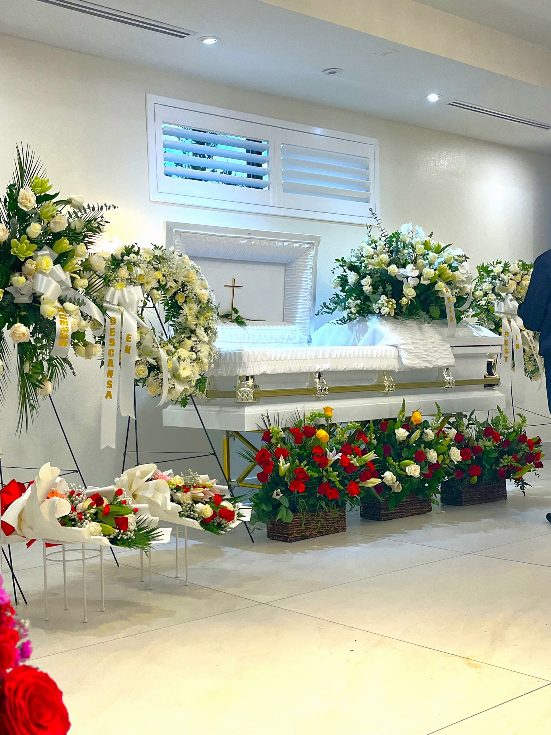 White casket surrounded by white, yellow, and red funeral flowers in a bright chapel