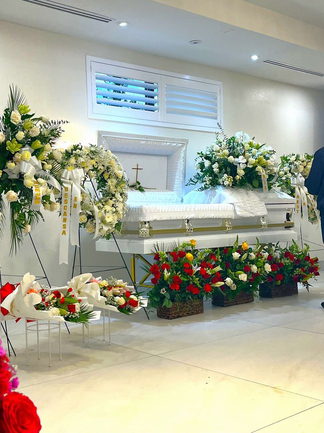 White casket surrounded by white, yellow, and red funeral flowers in a bright chapel