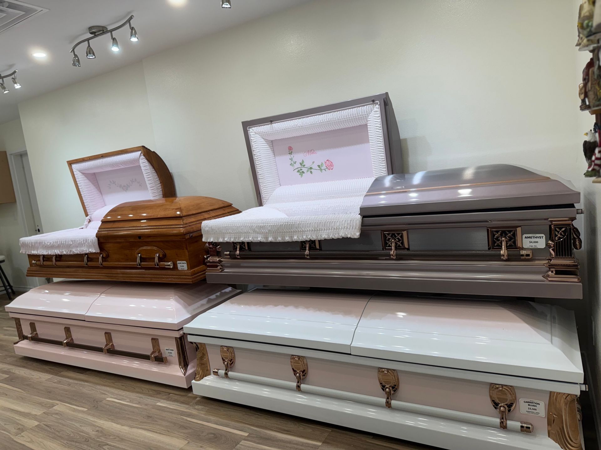 Display of several caskets in a funeral showroom, in white, brown, and silver finishes.