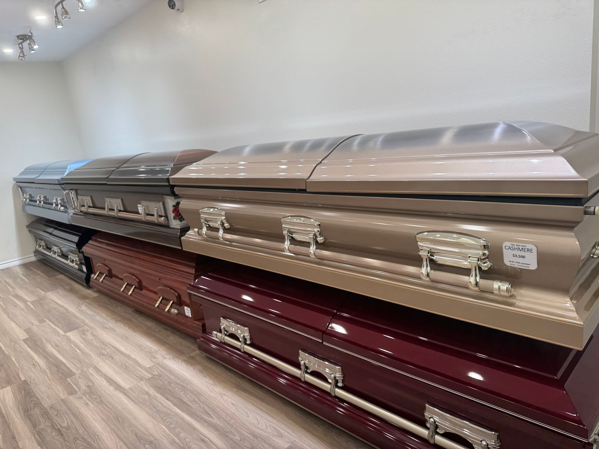 Three rows of glossy caskets in a showroom with wood flooring and white walls.
