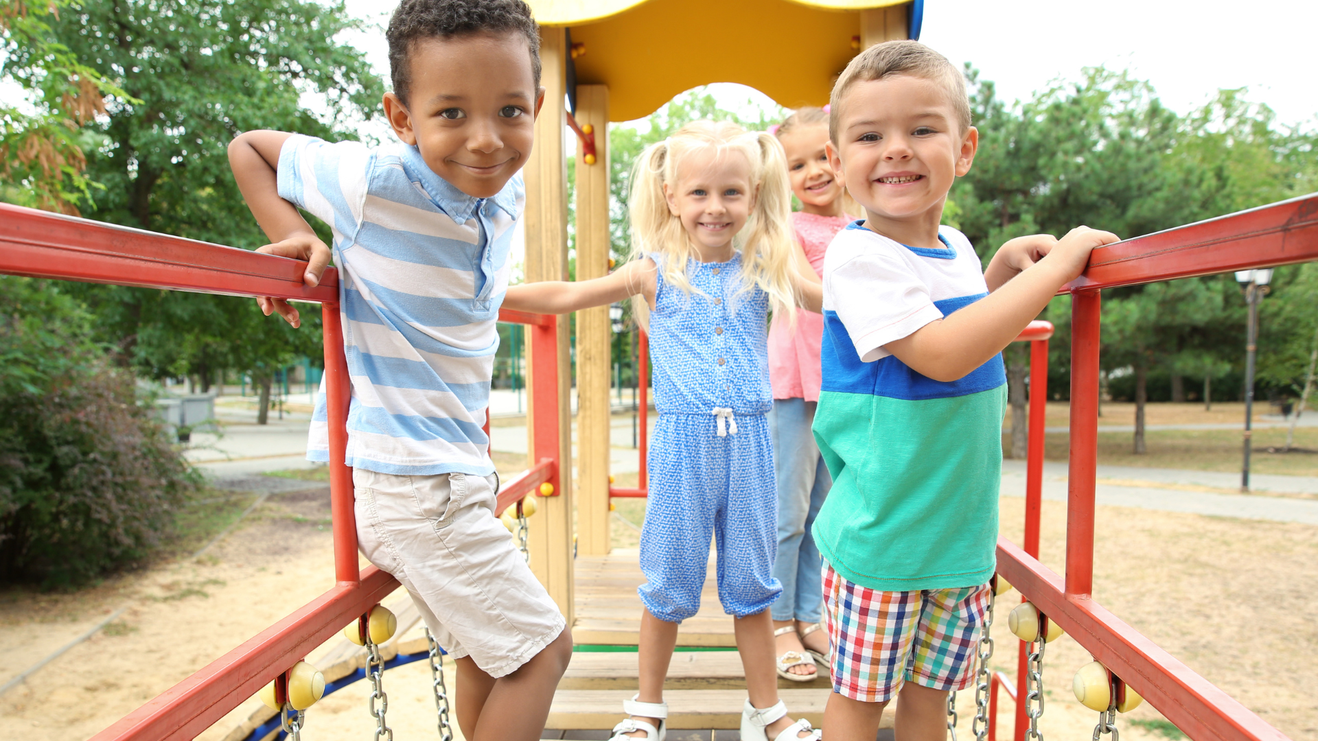 Three children smiling on a playground bridge with red rails, holding the bars outdoors.
