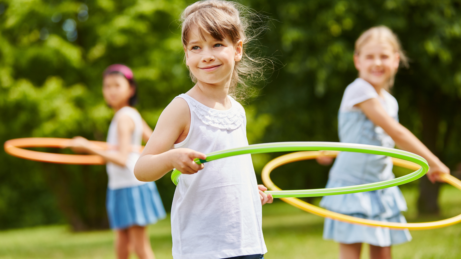 Child smiling while holding a hula hoop in a grassy park, with two others blurred in the background.