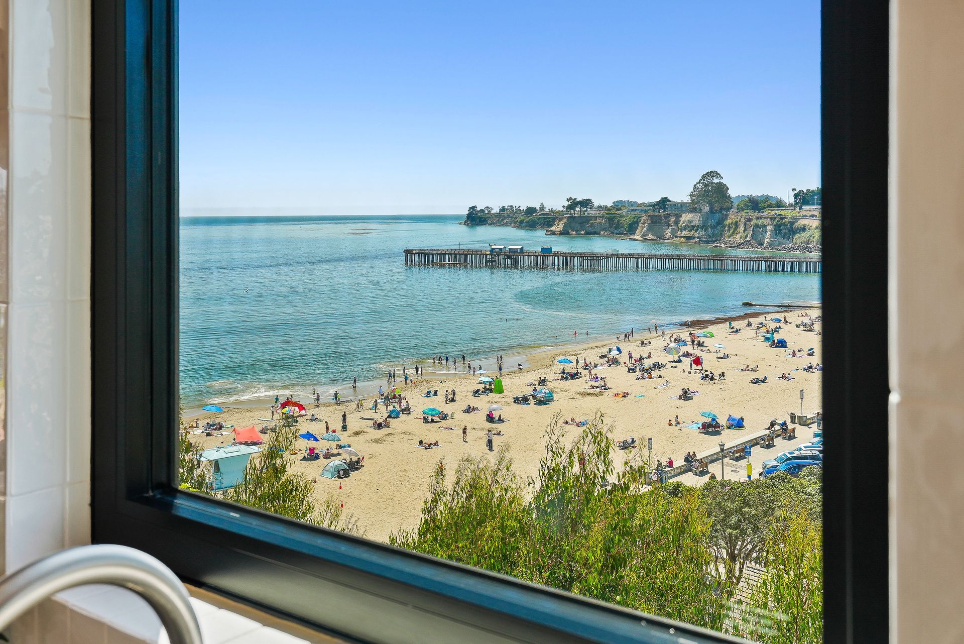Beach and calm blue sea viewed through a window, with a pier and umbrellas along the shore