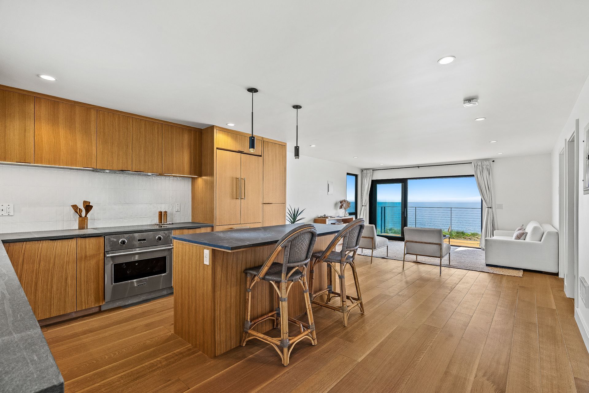 Modern open-plan kitchen and living room with wood floors, island seating, and ocean view through large windows