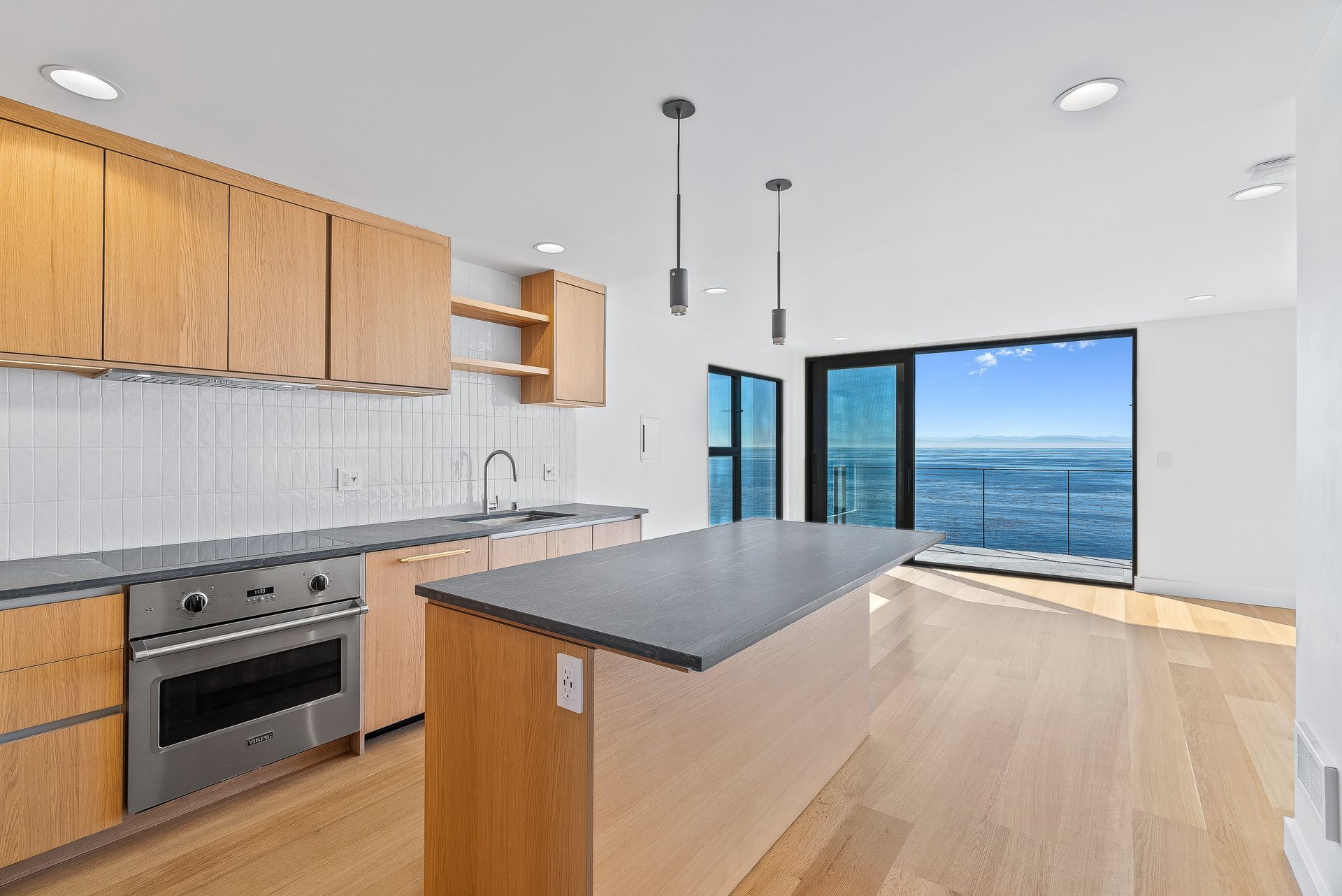 Modern kitchen with wood cabinets, black island, light flooring, and large windows overlooking water.