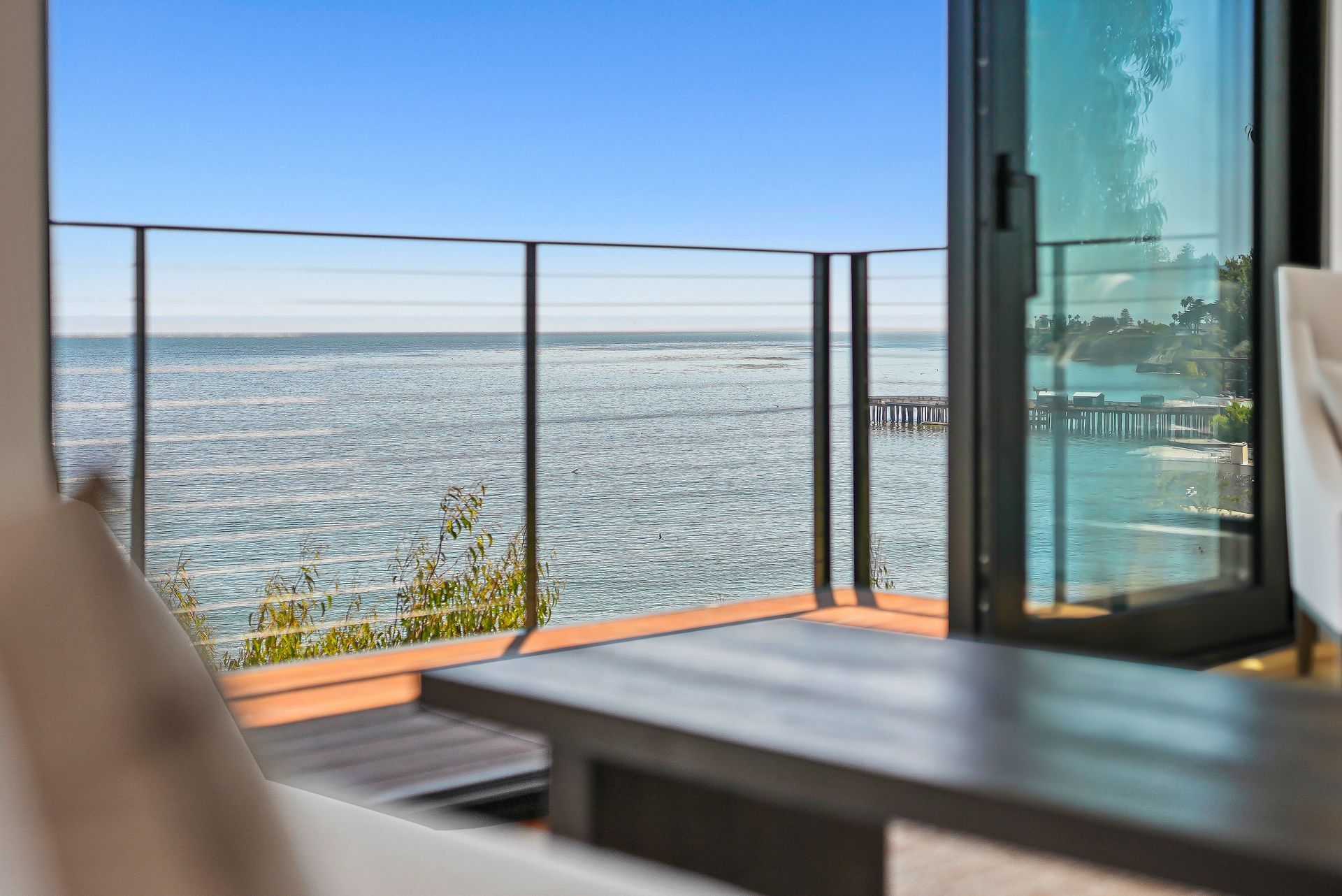 Seaside balcony with glass railing, wooden table, and ocean view under a clear blue sky