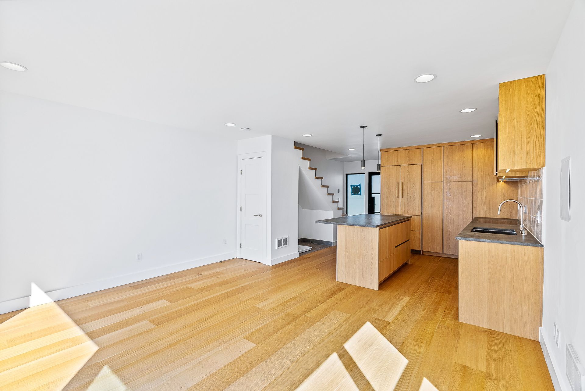 Empty modern open-plan apartment with light wood floors, white walls, kitchen cabinets, and a staircase