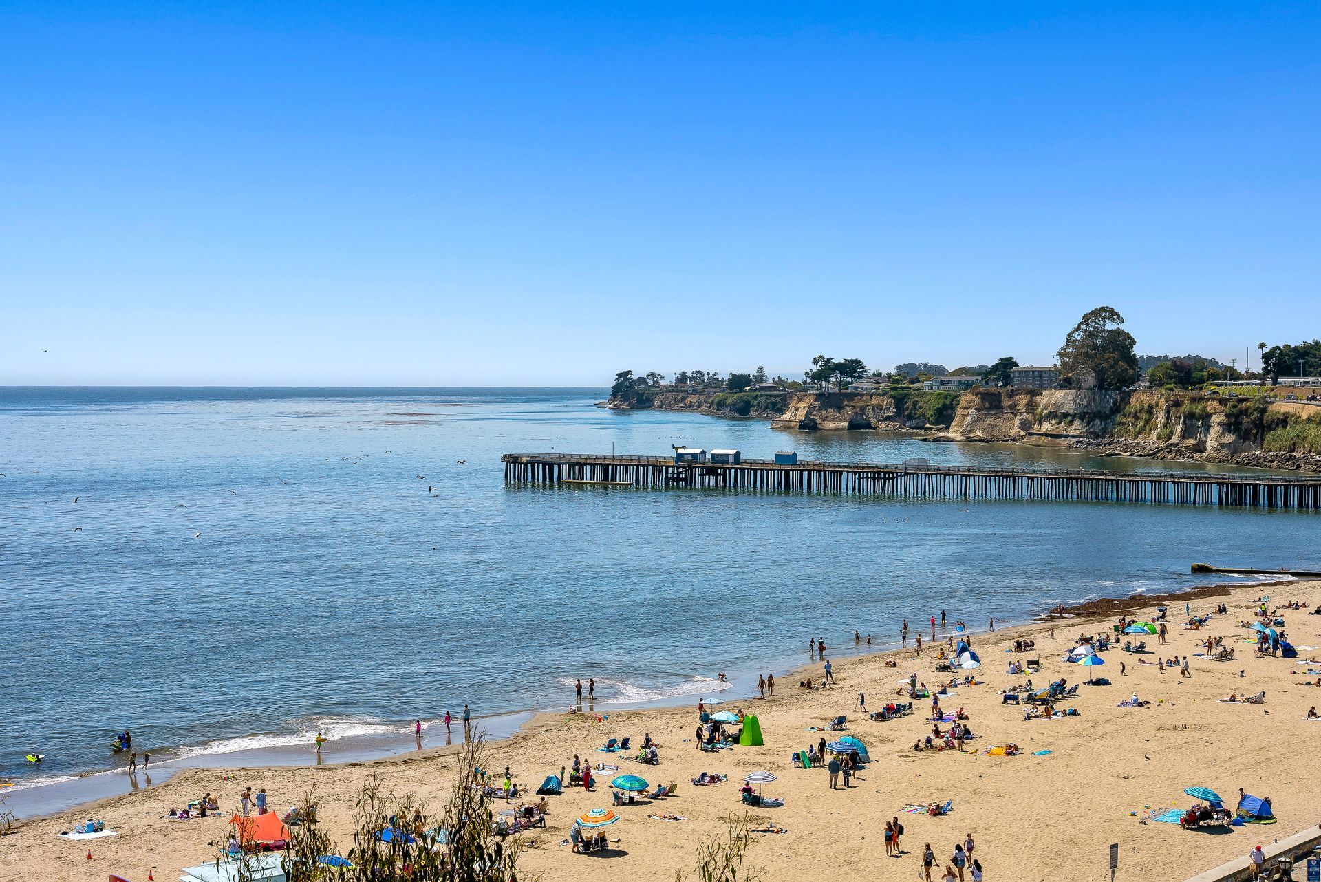 Sunny beach with scattered people, calm blue ocean, and a pier along the coast