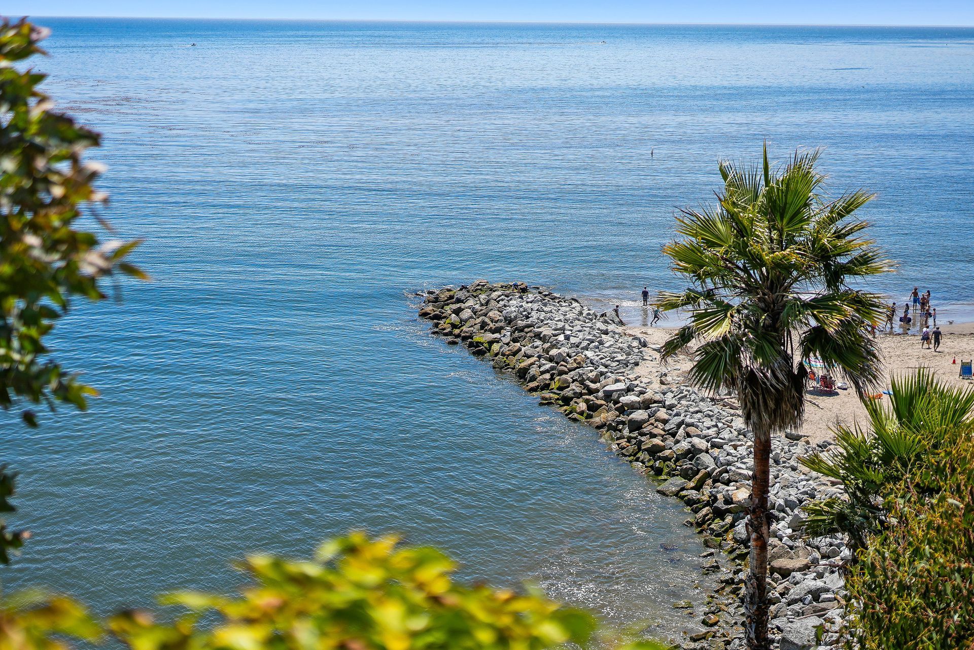 Rocky jetty extending into a calm blue sea, with palm trees and a sandy shoreline on a sunny day