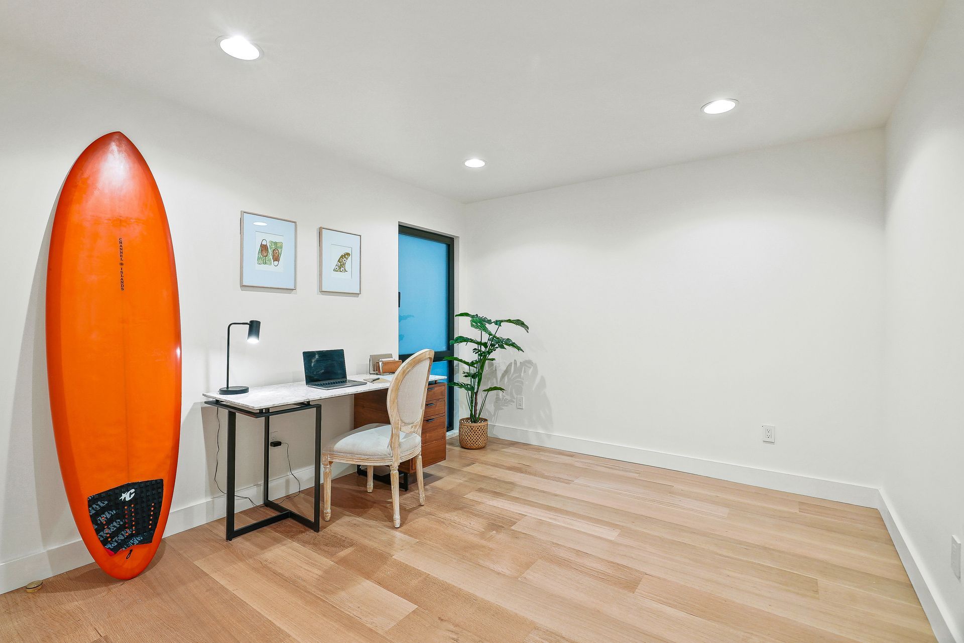 Bright minimalist room with desk, chair, plant, and an orange surfboard against the wall.