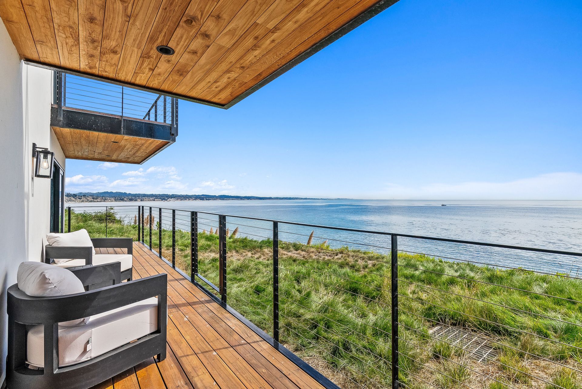 Covered wooden deck overlooking calm ocean, with glass railing and outdoor seating under a clear blue sky