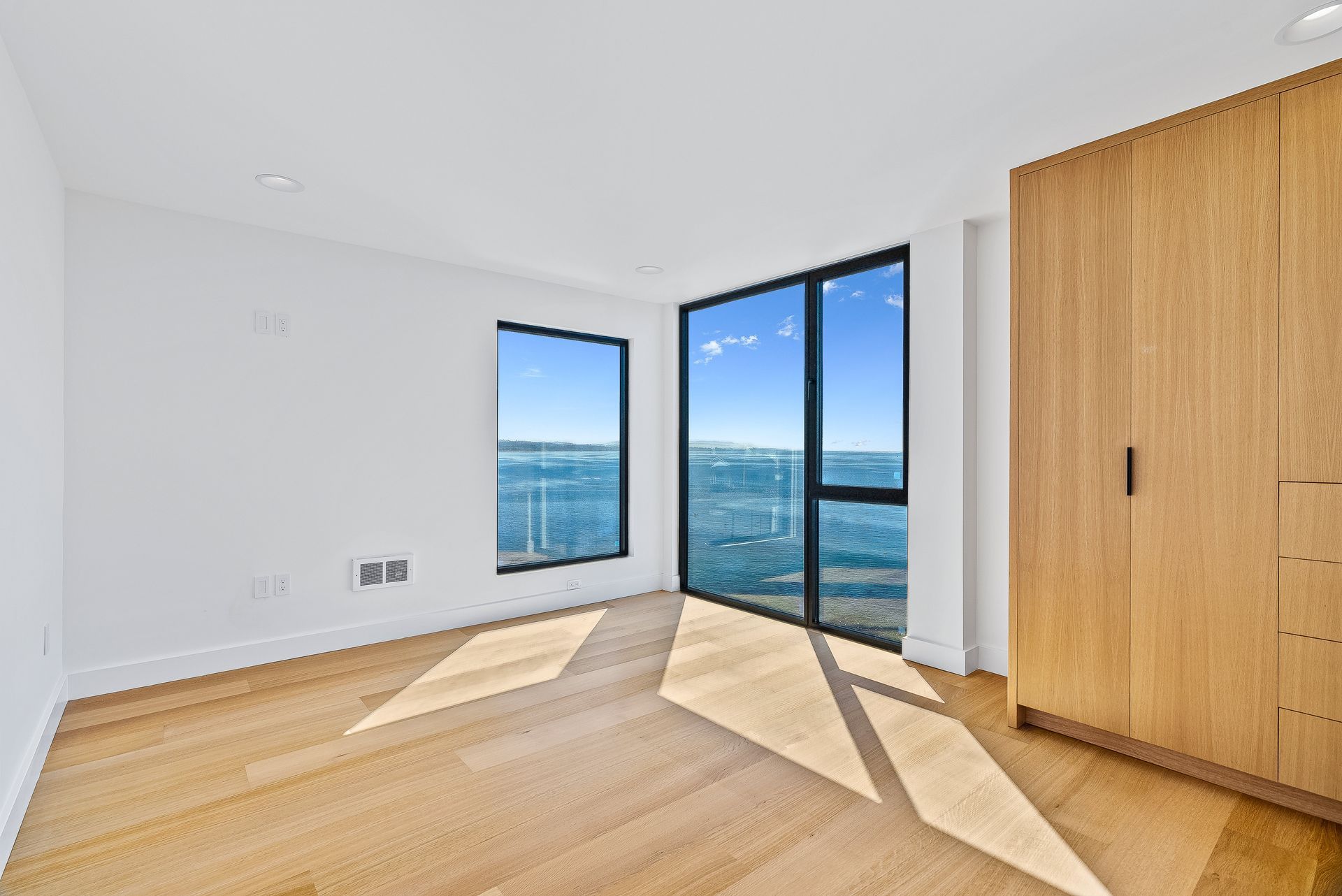 Bright empty room with wood floor, large windows, and ocean view in sunlight