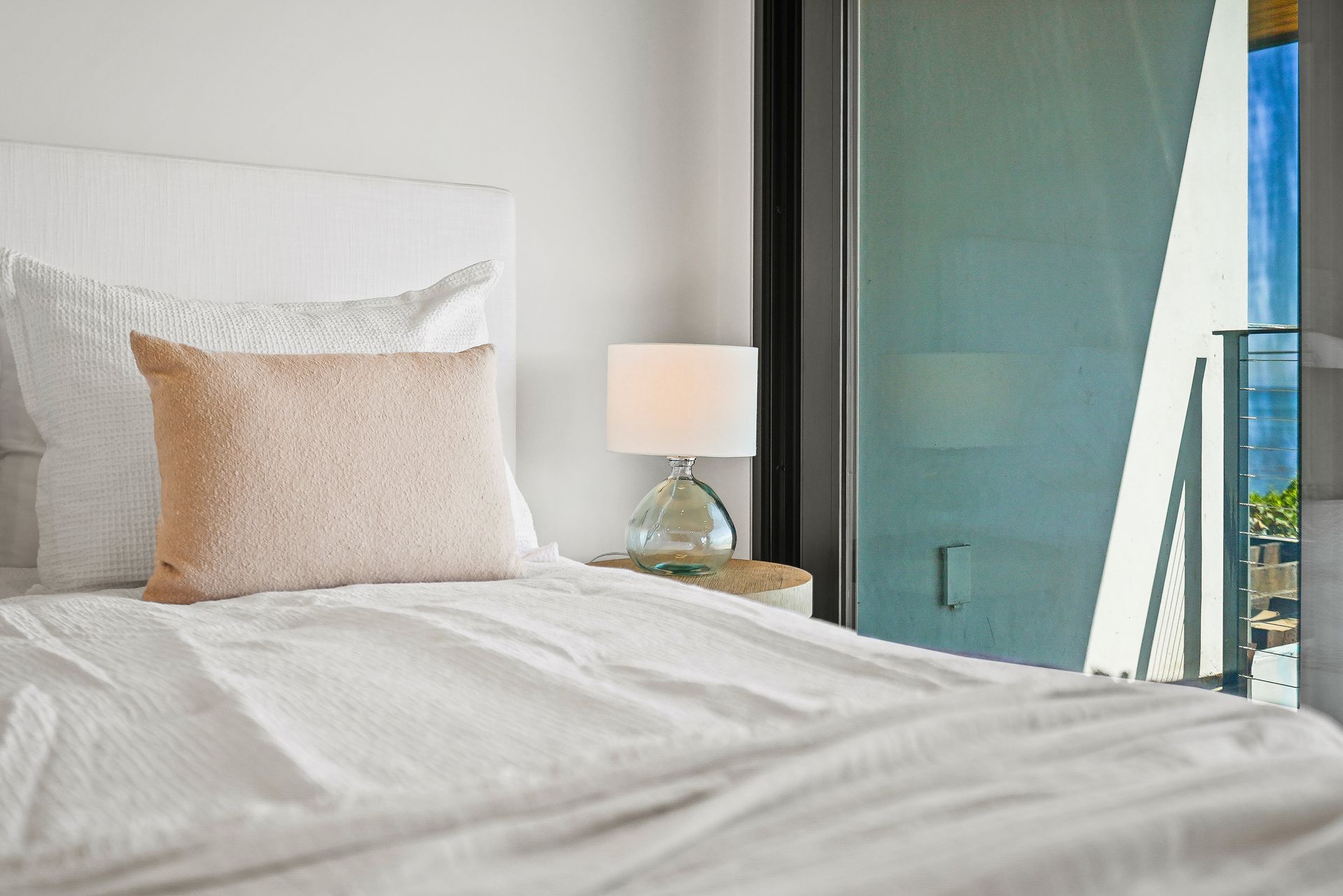 White bed with beige pillow beside a lamp and open balcony door overlooking the ocean
