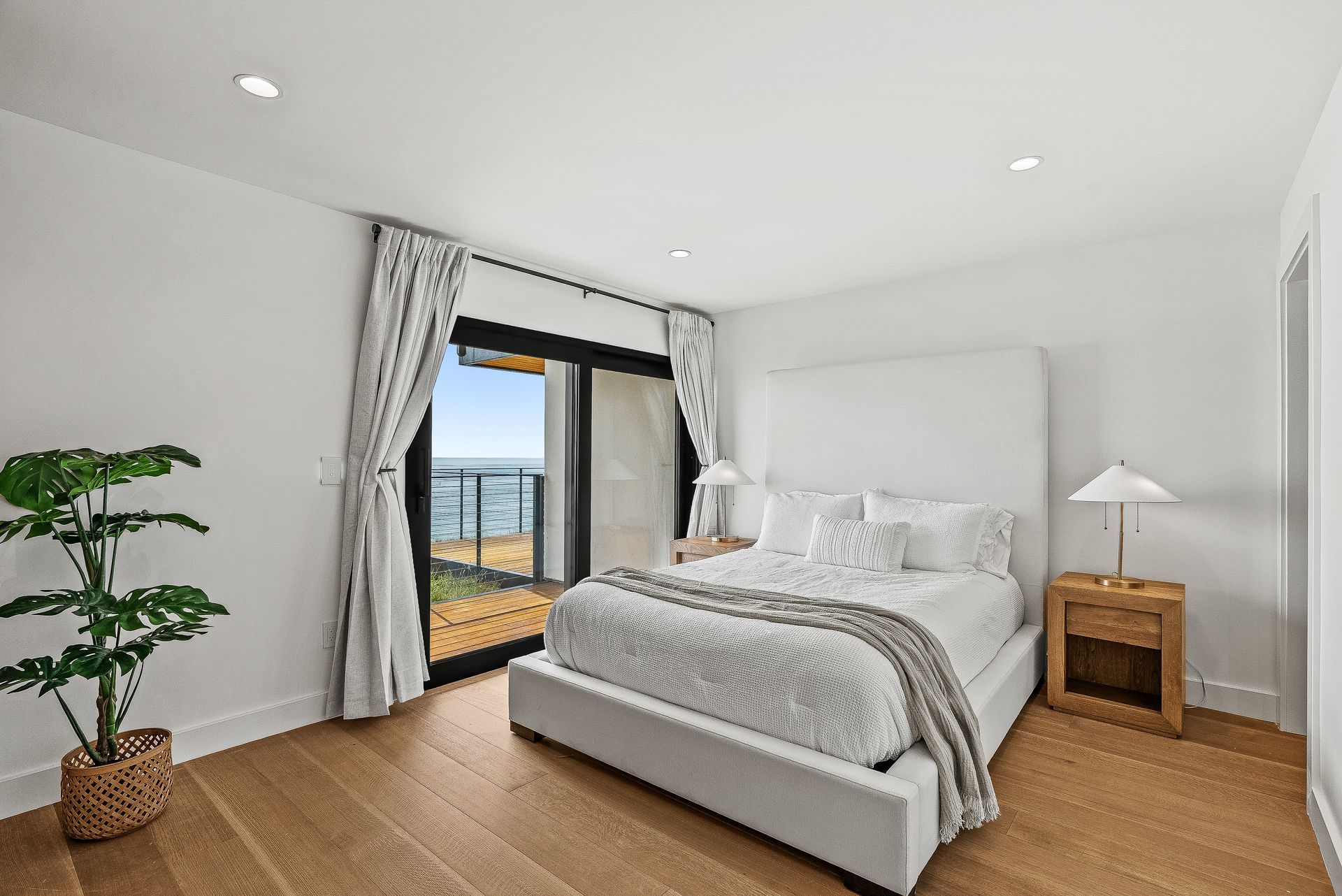 Bright modern bedroom with white bed, wood floor, potted plant, and balcony doors opening to the sea