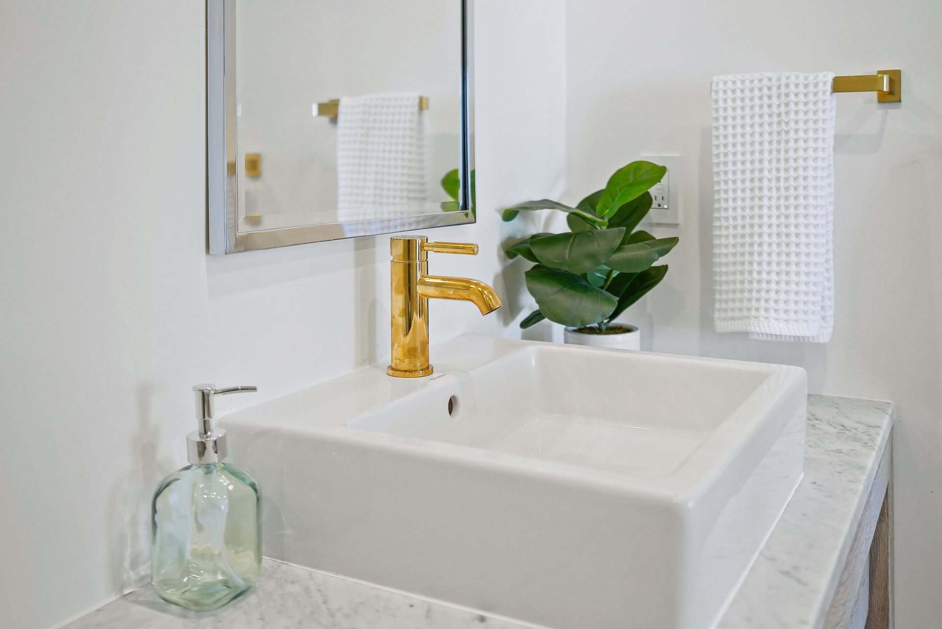 Bright bathroom sink with gold faucet, mirror, potted plant, and glass soap dispenser