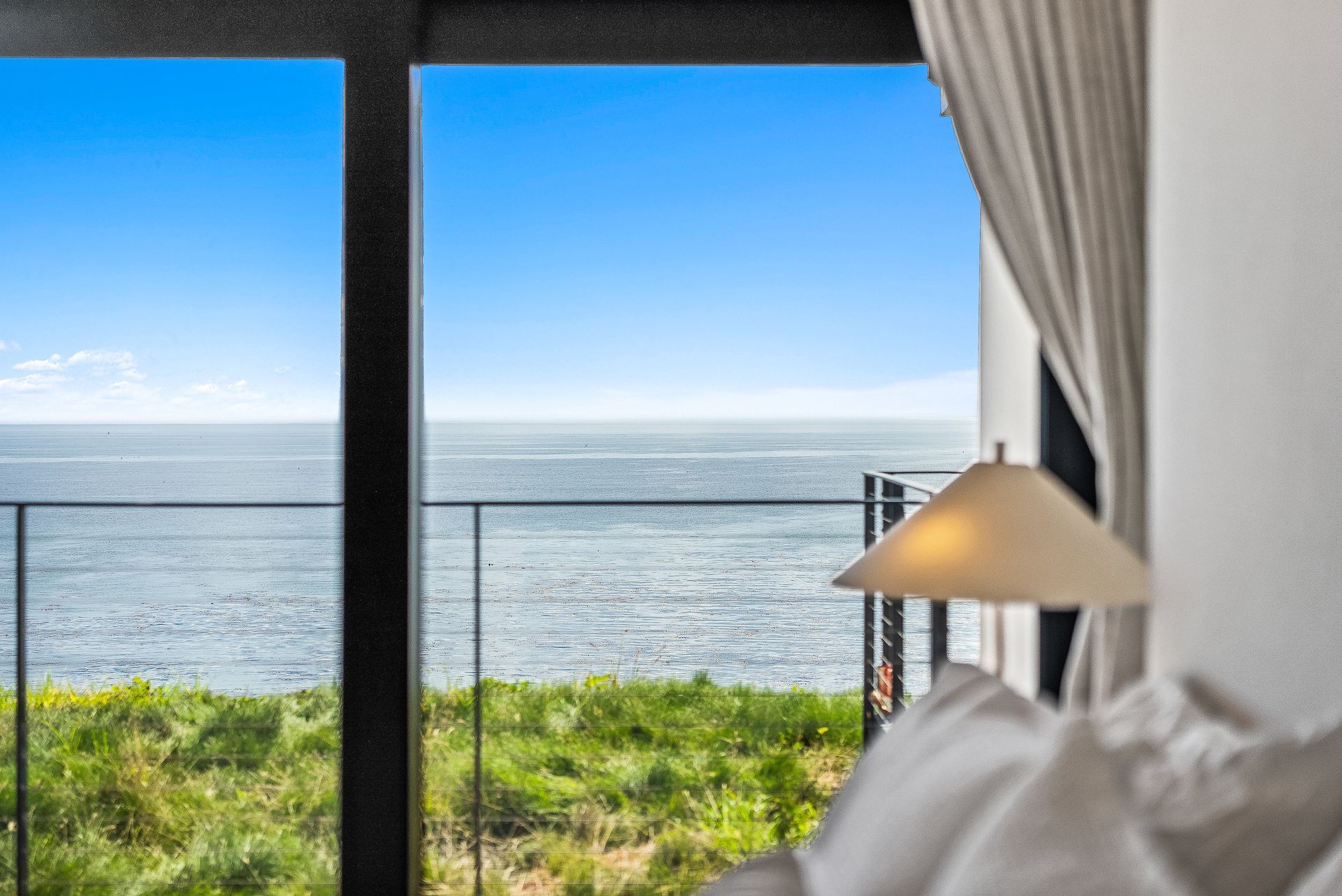 Ocean view through a window with a bed, lamp, and curtain in the foreground